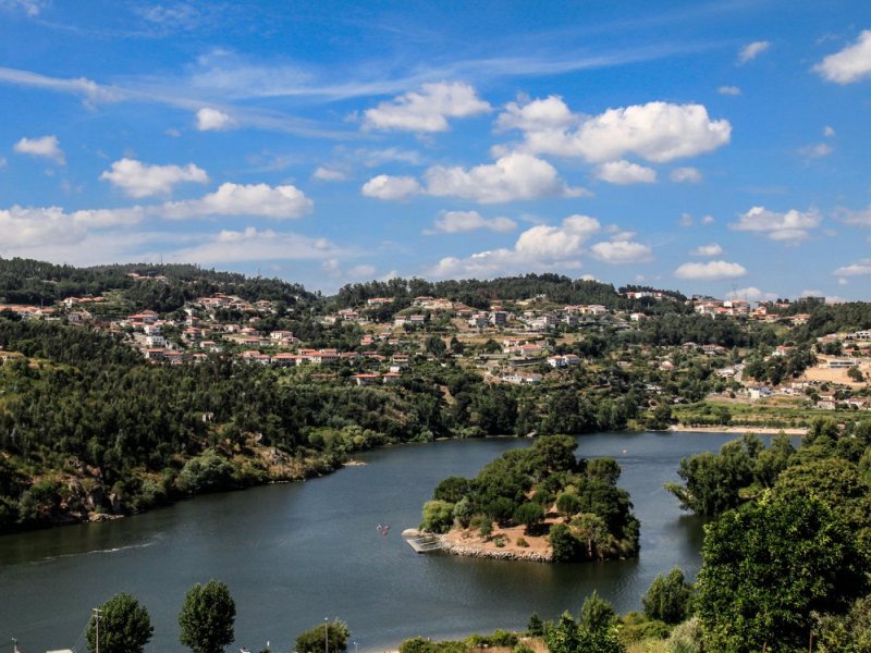 A picturesque landscape with a river, lush hills, and scattered houses under a blue sky with clouds.
