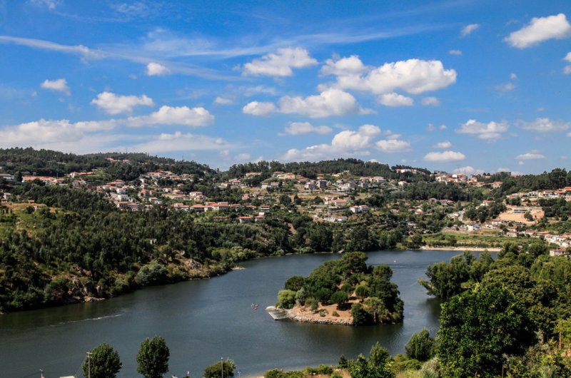 A picturesque landscape with a river, lush hills, and scattered houses under a blue sky with clouds.