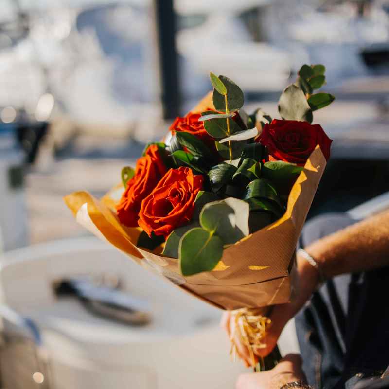 A person holds a bouquet of red roses wrapped in paper near a boat dock.