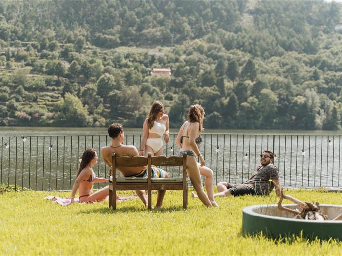 Group of people relaxing by a lakeside, with hills and trees in the background.