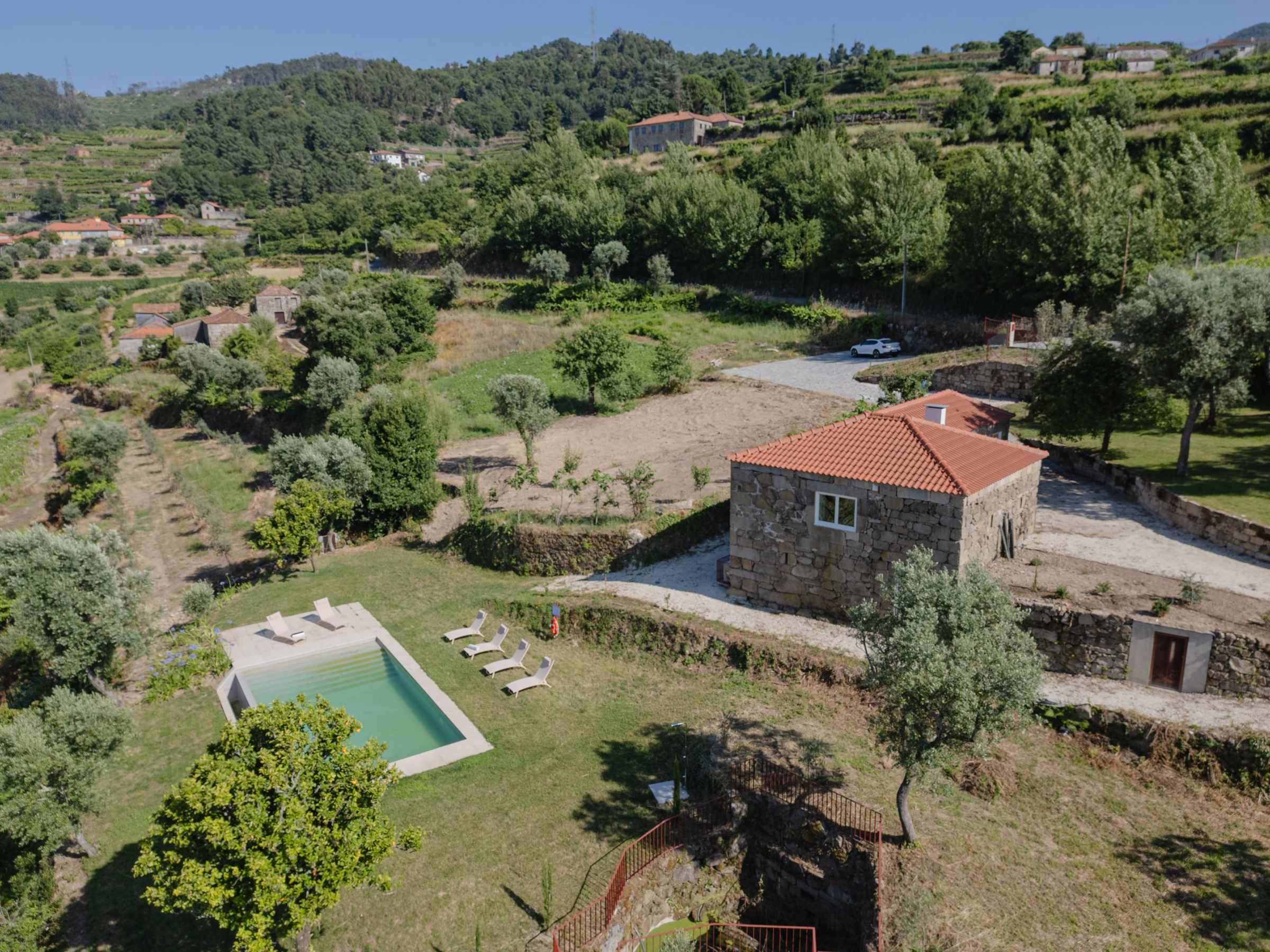Aerial view of a house with a red roof, a pool, and surrounding greenery in a rural landscape.