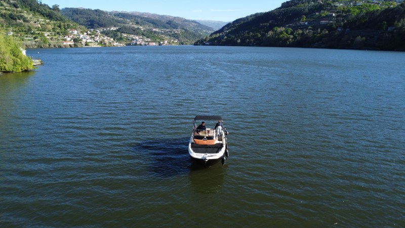 Small boat with people in the middle of a wide river, surrounded by hills and distant town.