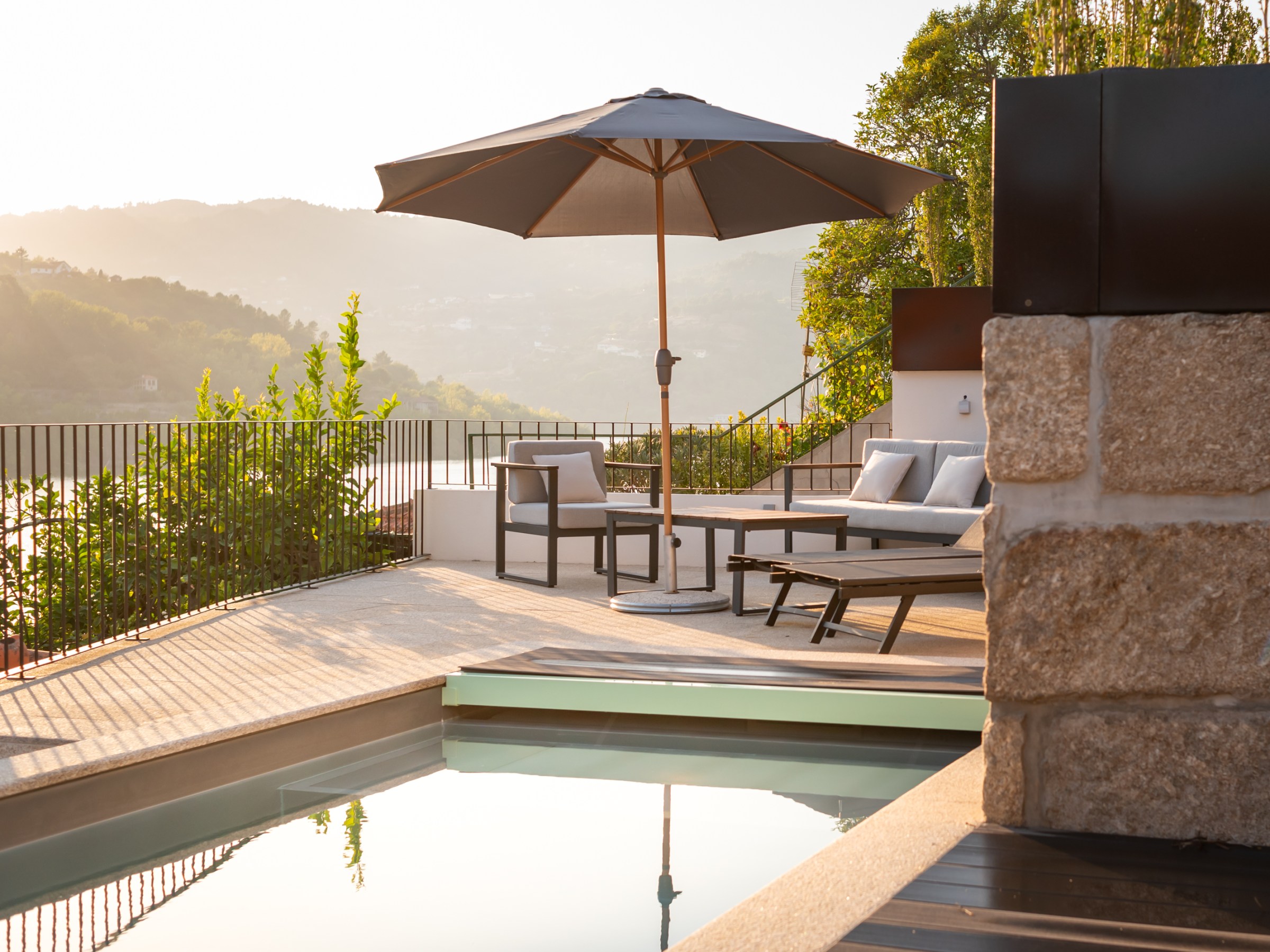 Sunlit patio with chairs, umbrella, and pool overlooking a scenic hillside.