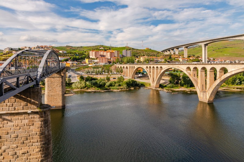 Two bridges crossing a river with a town and hills in the background under a cloudy sky.