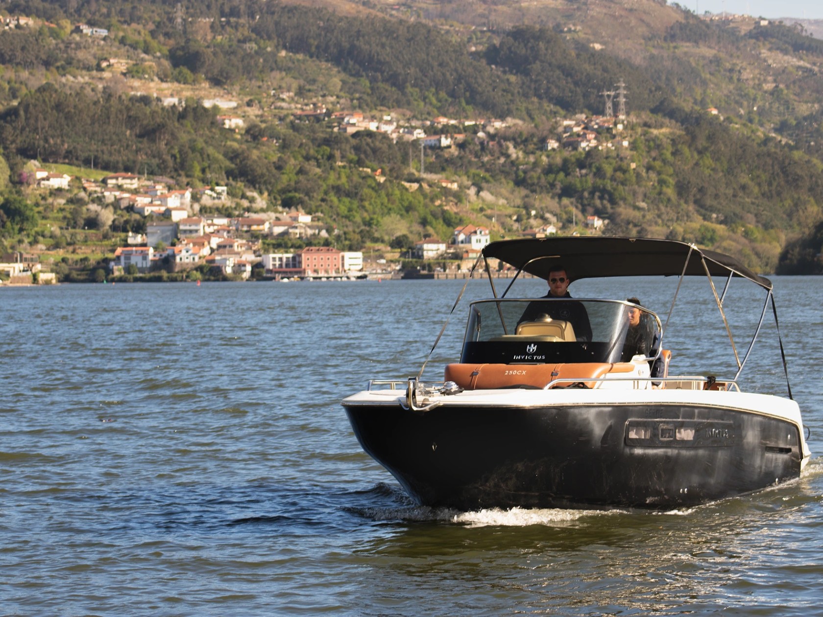 Small motorboat on river with person, hills and houses in background.
