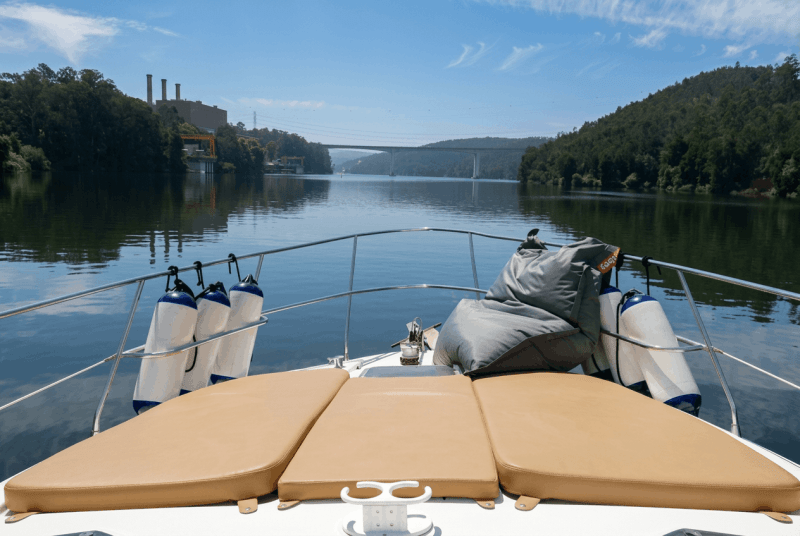View from a boat deck with cushions, calm river, trees, and distant bridge.