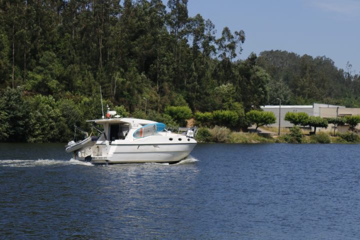White boat cruising on a calm river with trees and buildings in the background.