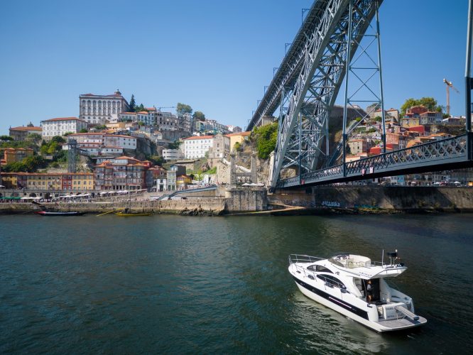 Boat on river beneath large metal bridge, colorful hillside buildings in background.