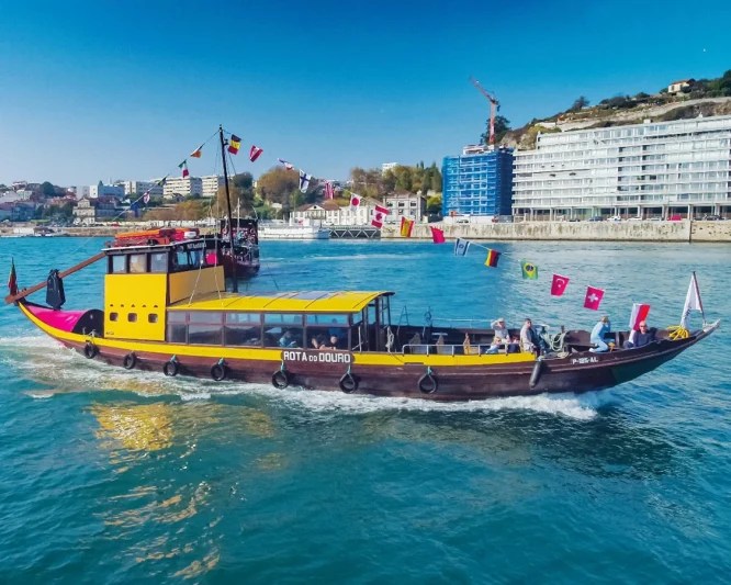 Wooden tour boat on a river with flags, city buildings in the background.