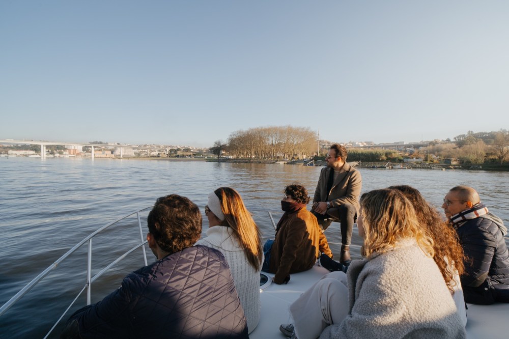 Group of people sitting aboard a boat on the Douro River in late afternoon, with golden light and the city of Porto in the background
