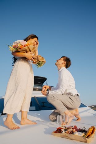 Man proposing on a boat, holding ring, woman smiling with flowers.