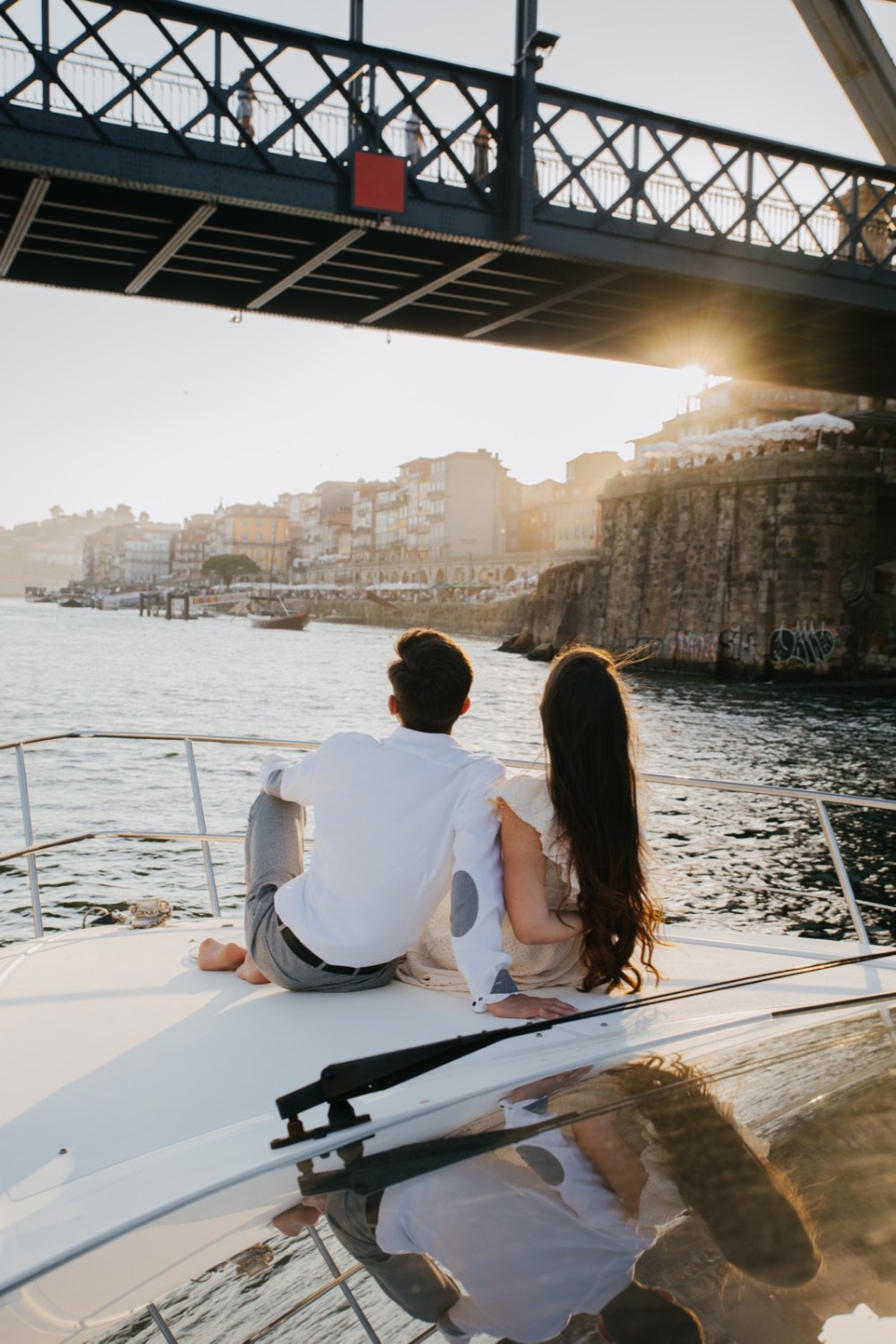 Couple sitting on a boat, looking at a bridge with sunset in the background.