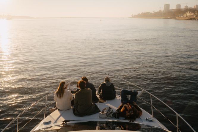 Four people sitting on a boat deck, looking at a calm body of water during sunset.
